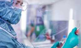 The picture shows a section of a laboratory worker in a clean room suit working on a sample from the right side.