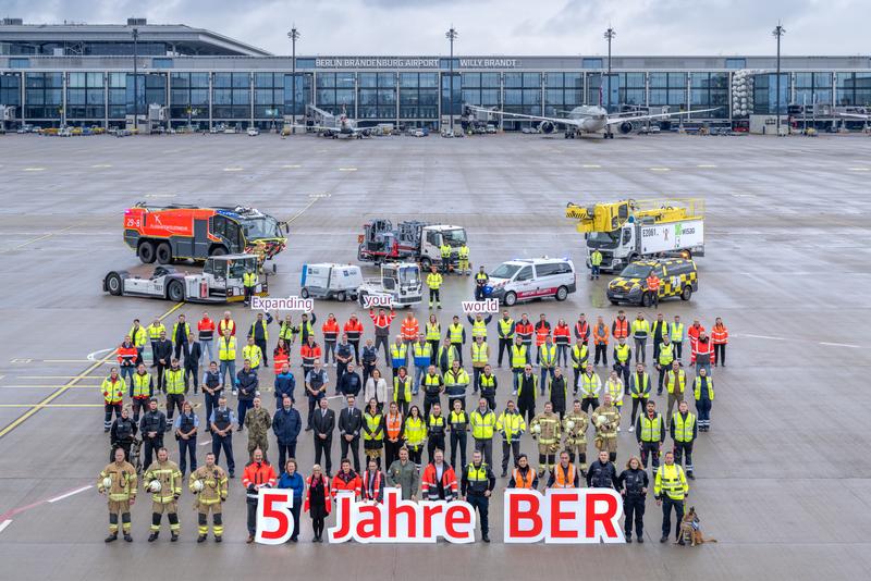 To mark BER’s anniversary, many employees wearing safety vests stand on the apron holding up symbolic signs, with the airport and several aircraft visible in the background.