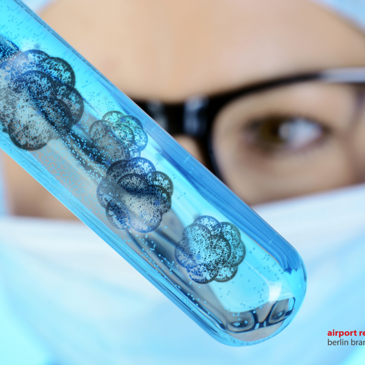 In the foreground is a test tube with cells, in the background a slightly blurred face of a researcher with a mask and glasses looking at the test tube.