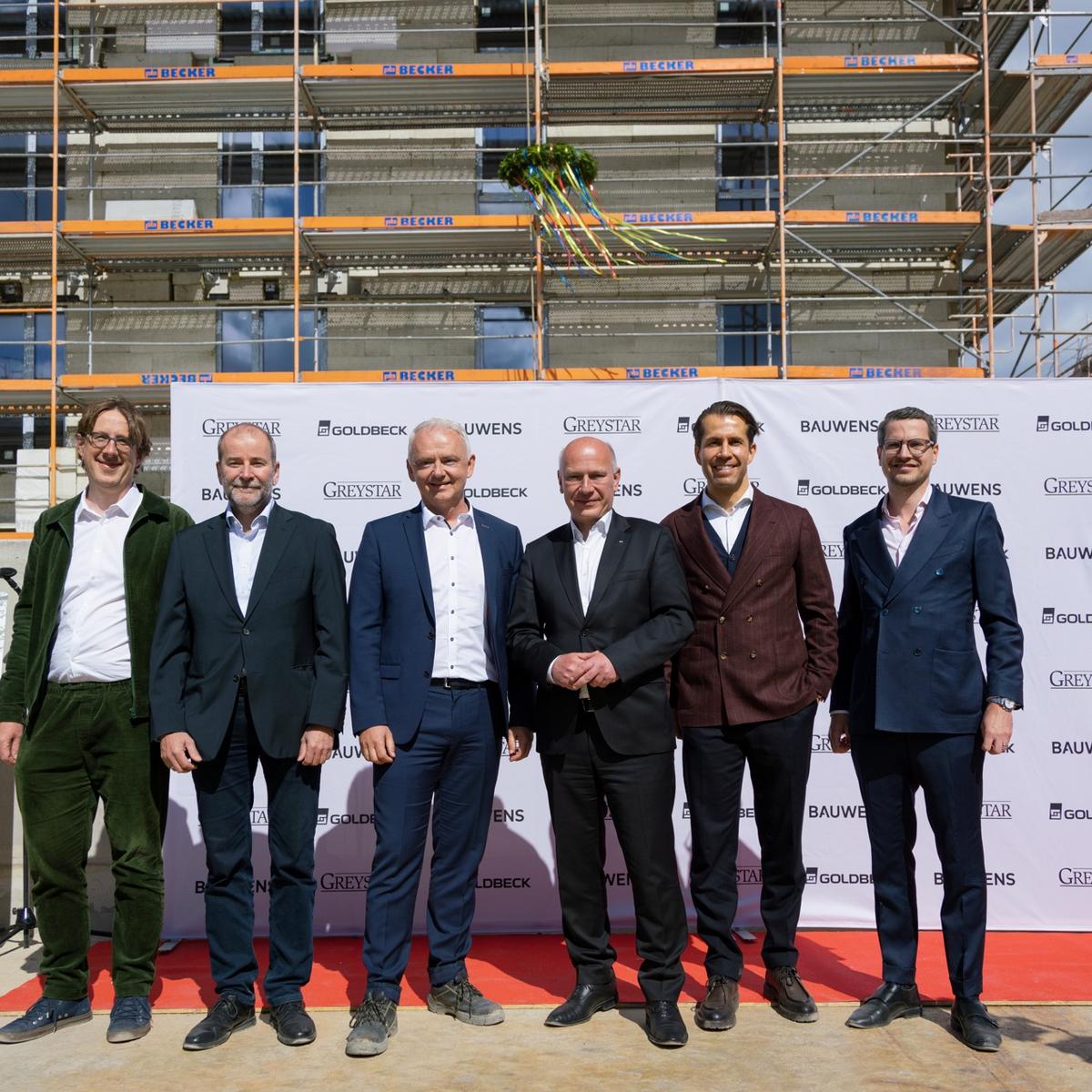 The following people are standing together in front of a logo banner at the Greenpark topping-out ceremony in Berlin: District Councillor Jochen Biedermann, Udo Teschner (Berlinovo), Robert Wall (Goldbeck), Governing Mayor Kai Wegner, Clemens Stahr (Bauwens), and Maximilian Herbst (Greystar).