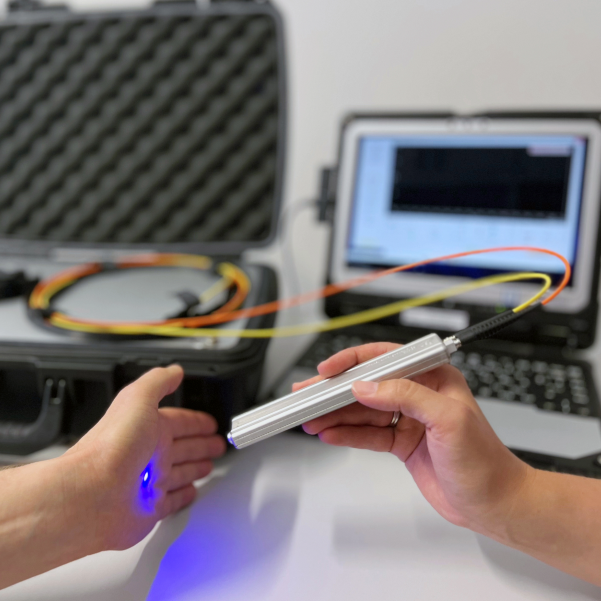 mage shows two hands holding an optical device in front of a measuring computer. The device generates a blue dot of light on the skin and uses it to measure certain values in the skin.