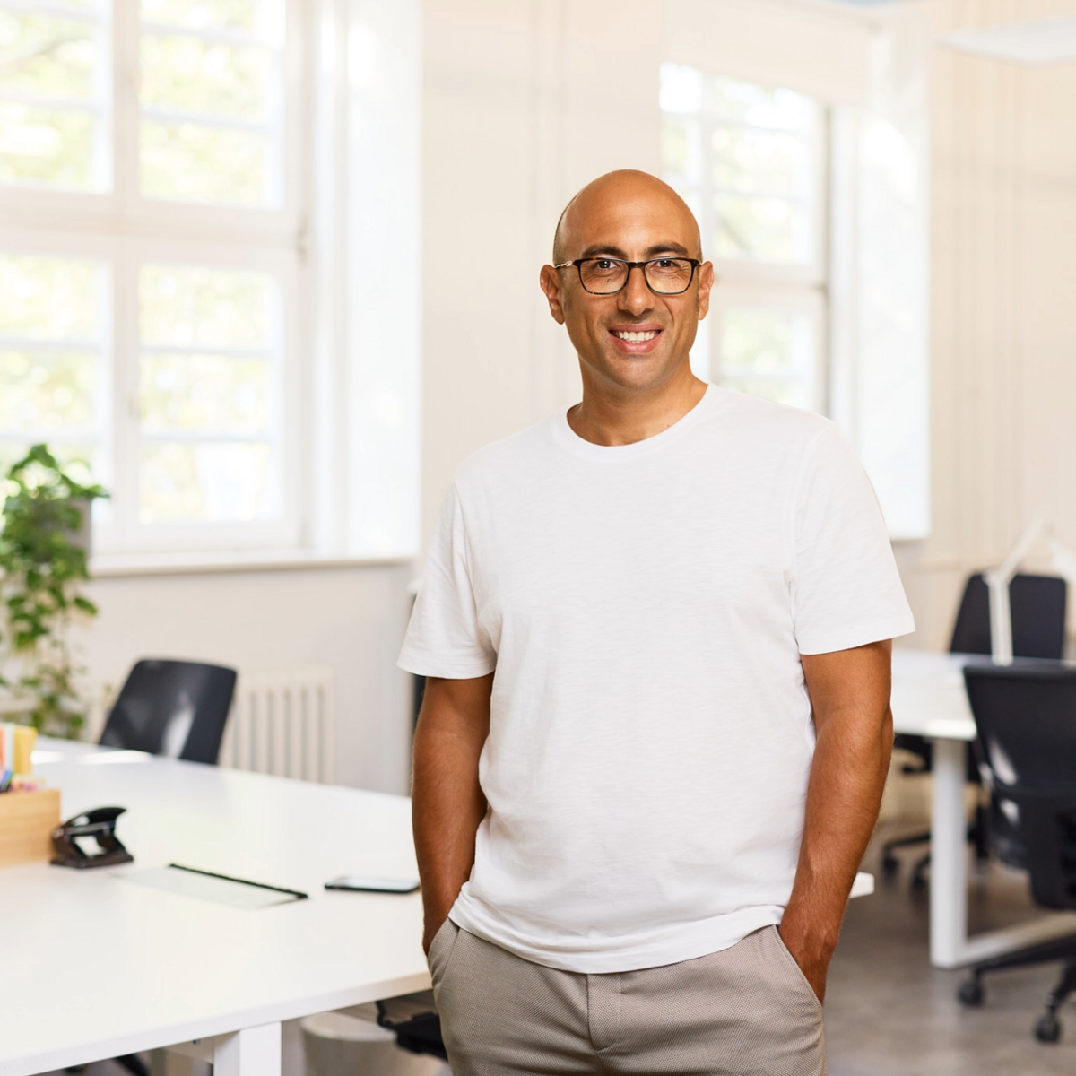 A man with a bald head, wearing glasses and a white T-shirt is standing in front of an office backdrop.