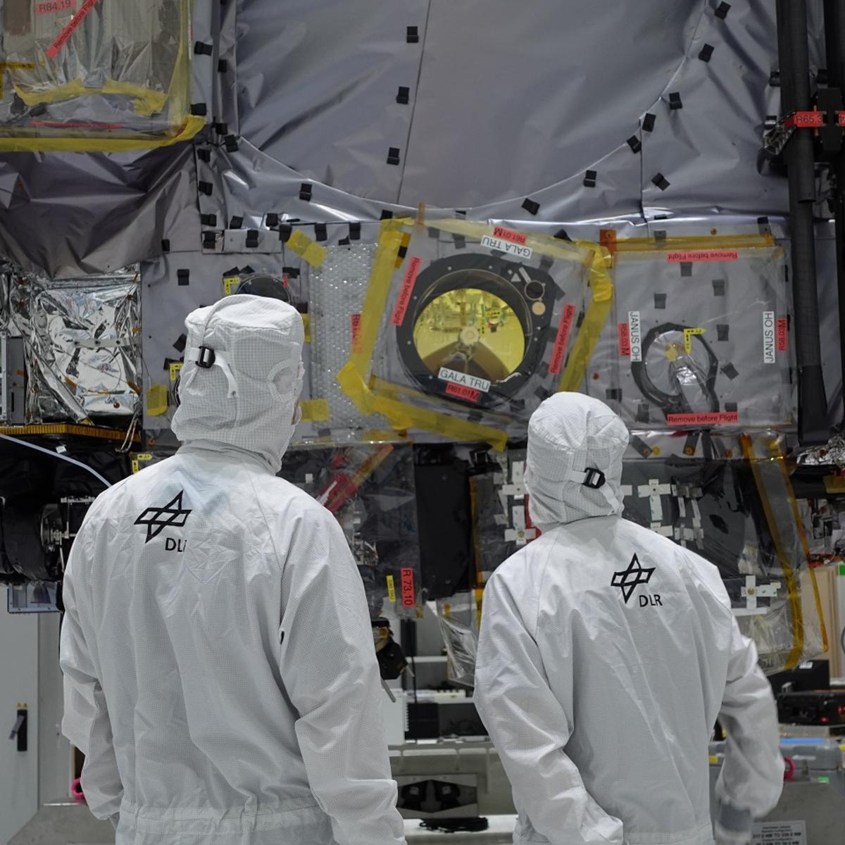 2 people in white protective suits are standing in front of the optical camera and laser altimeter for the ESA mission JUICE.