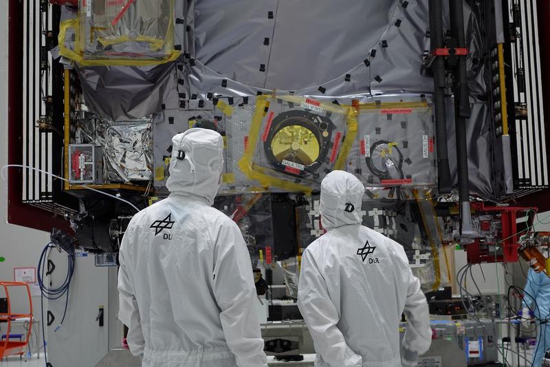 2 people in white protective suits are standing in front of the optical camera and laser altimeter for the ESA mission JUICE.