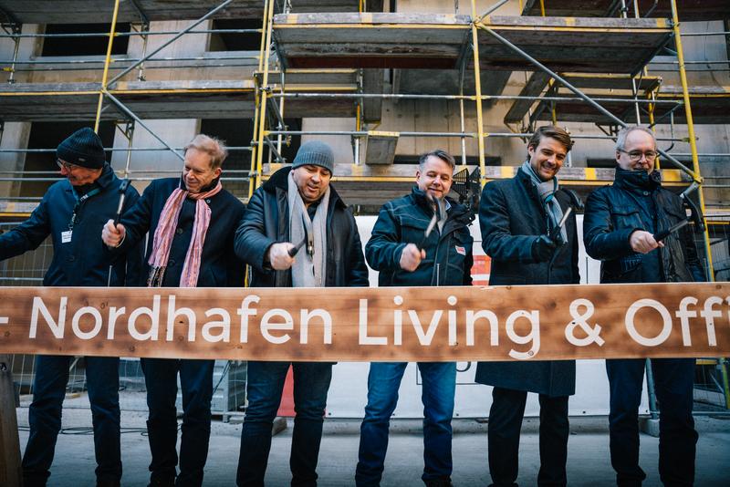 Several people participate in the ceremonial nail driving into a beam branded with Nordhafen Living & Office.