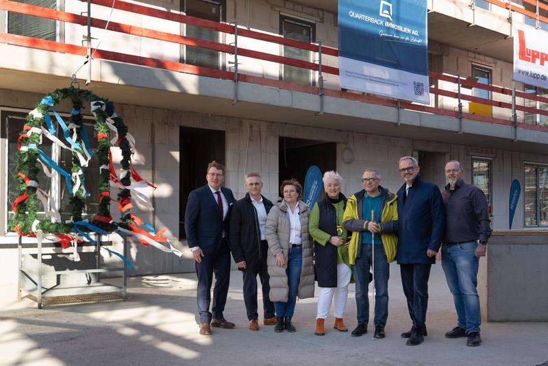 In front of the residential quarter at the topping-out ceremony, the following persons are standing side by side (from left to right): Steffen Hanke, Branch Manager Potsdam, QUARTERBACK Immobilien AG; Michael Fuchs, Managing Director, Adolf Lupp GmbH; Stefanie Löwe-Koch, Head of Office Management Office East/West & Residential, HIH Group; Michaela Wiezorek, Mayor of Königs Wusterhausen; Georg Hanke, Local Representative, District of Königs Wusterhausen; Henrik Thomsen, Board Member, QUARTERBACK Immobilien AG; Nils Witschonke, Senior Construction Manager, Adolf Lupp GmbH.