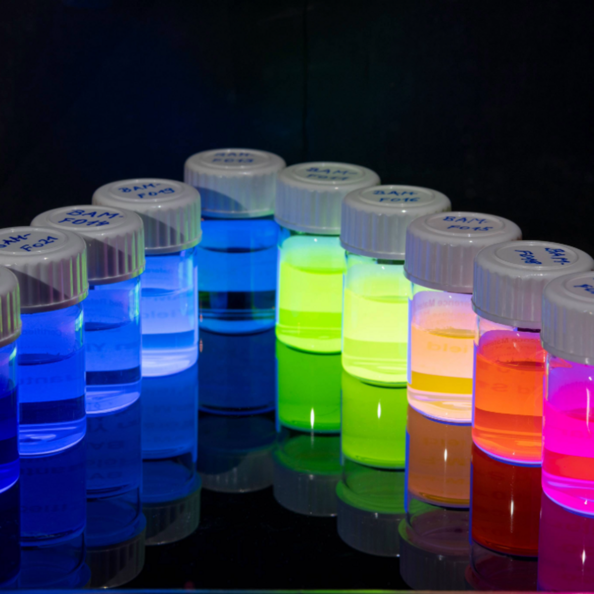 The image shows 12 small containers sorted by color, containing glowing nanoparticles.