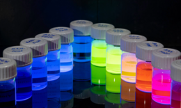 The image shows 12 small containers sorted by color, containing glowing nanoparticles.
