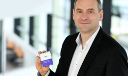The image shows a smiling man holding a small test kit box and showing it to the camera. The background is blurred.
