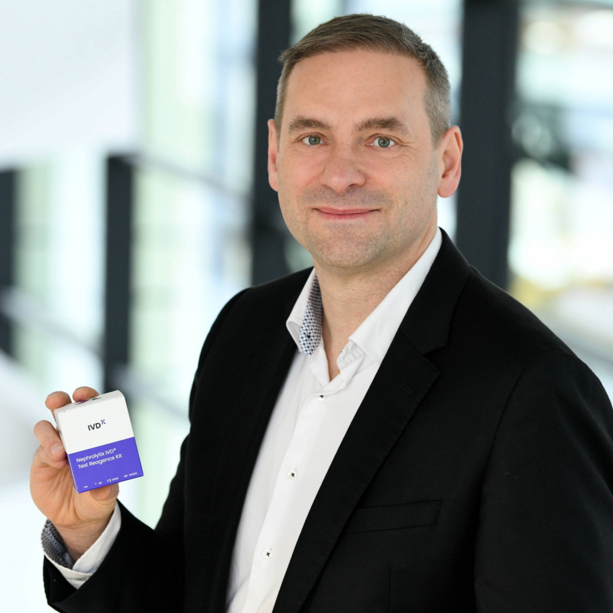 The image shows a smiling man holding a small test kit box and showing it to the camera. The background is blurred.