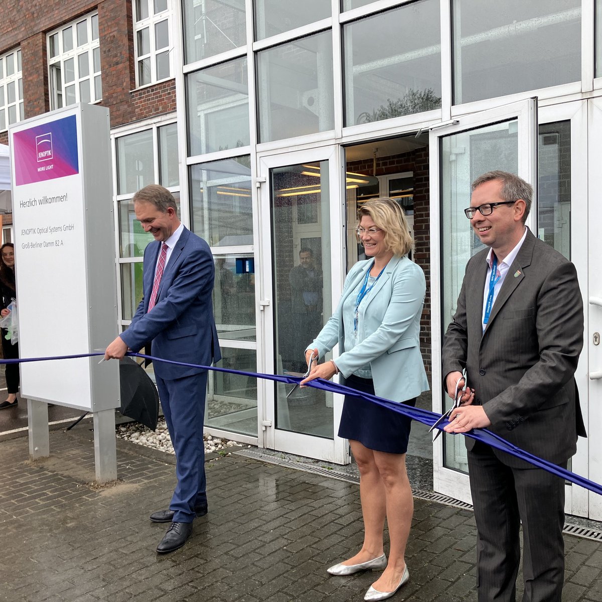 The picture shows a woman and two men people ceremoniously cutting a ribbon to open a new company location.
